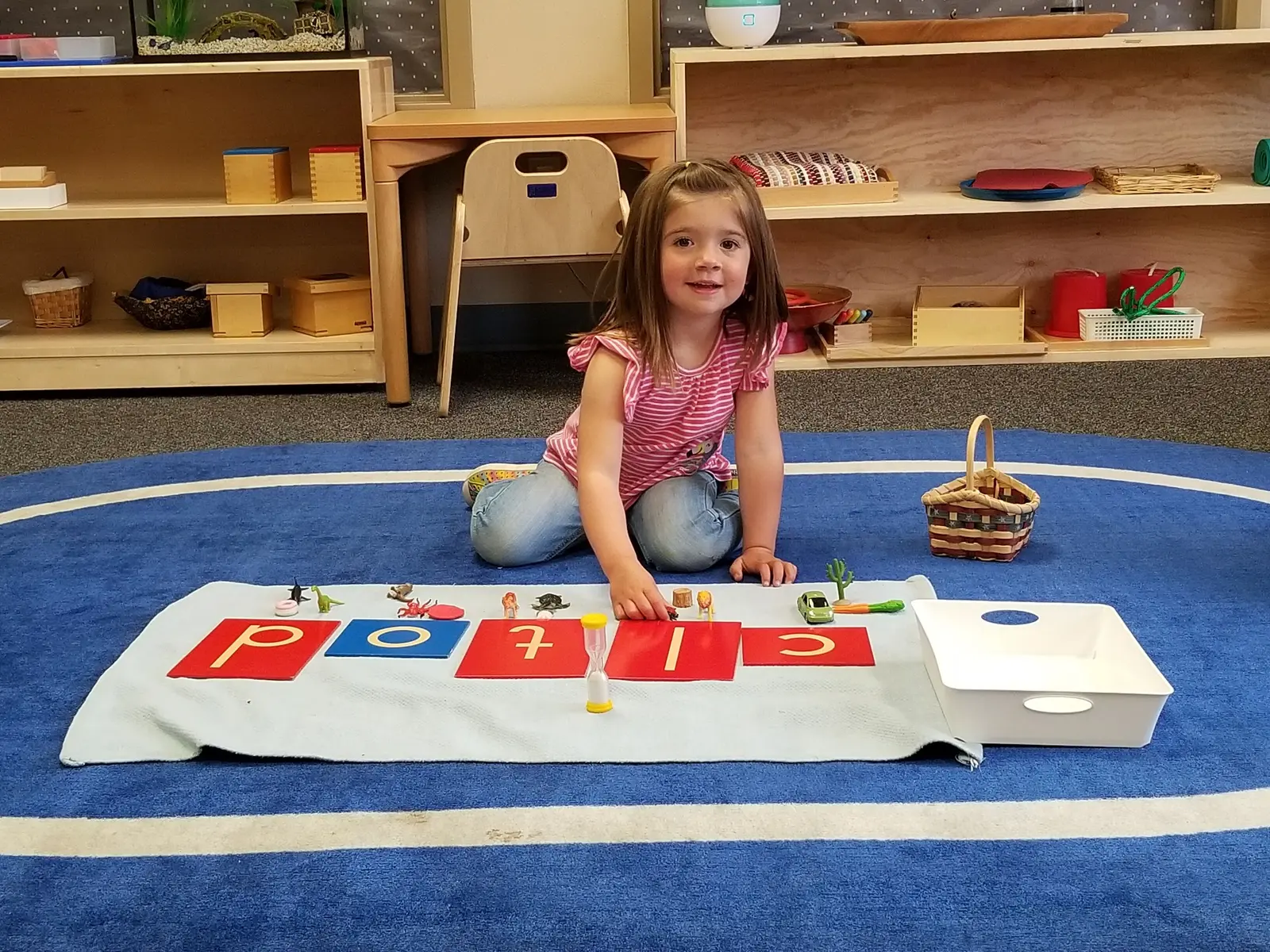 A young girl sits on the floor in a classroom playing with letter tiles and toy figures.