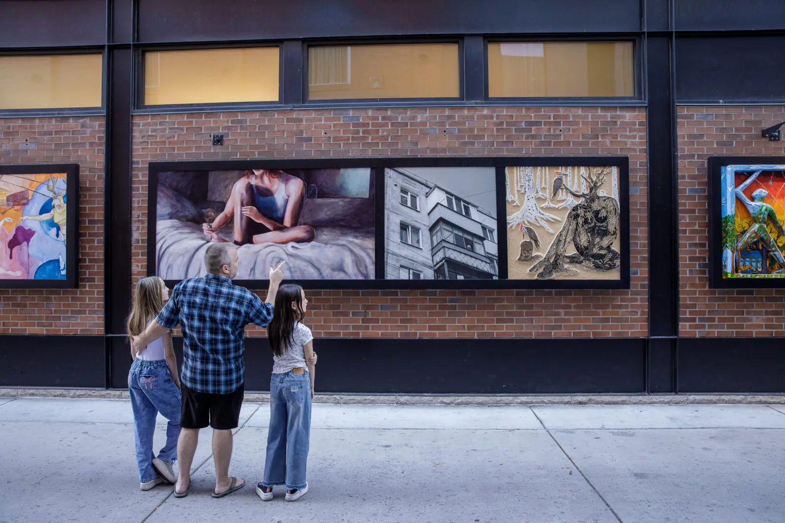 A man and two girls stand in front of a series of paintings displayed on a brick wall.