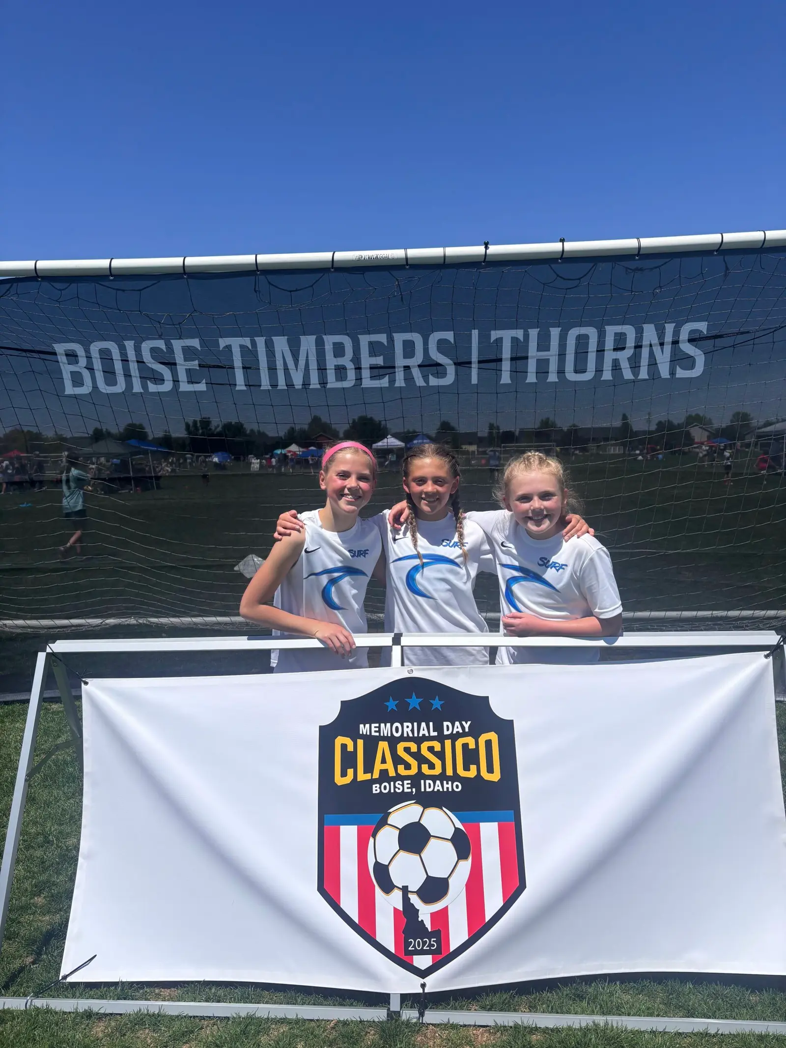 Three young soccer players stand together in front of a tournament banner.