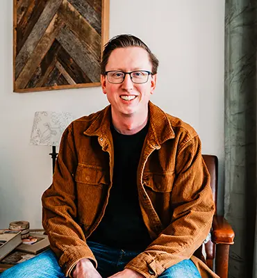 A man wearing glasses and a brown jacket smiles while sitting in a room.