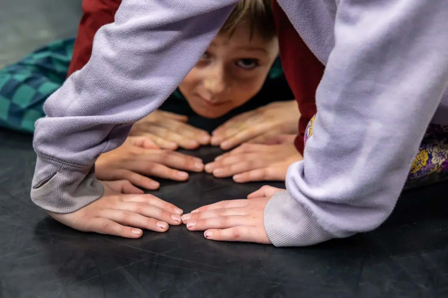 Several children are reaching their hands toward the center on a dark surface while one looks through their arms.