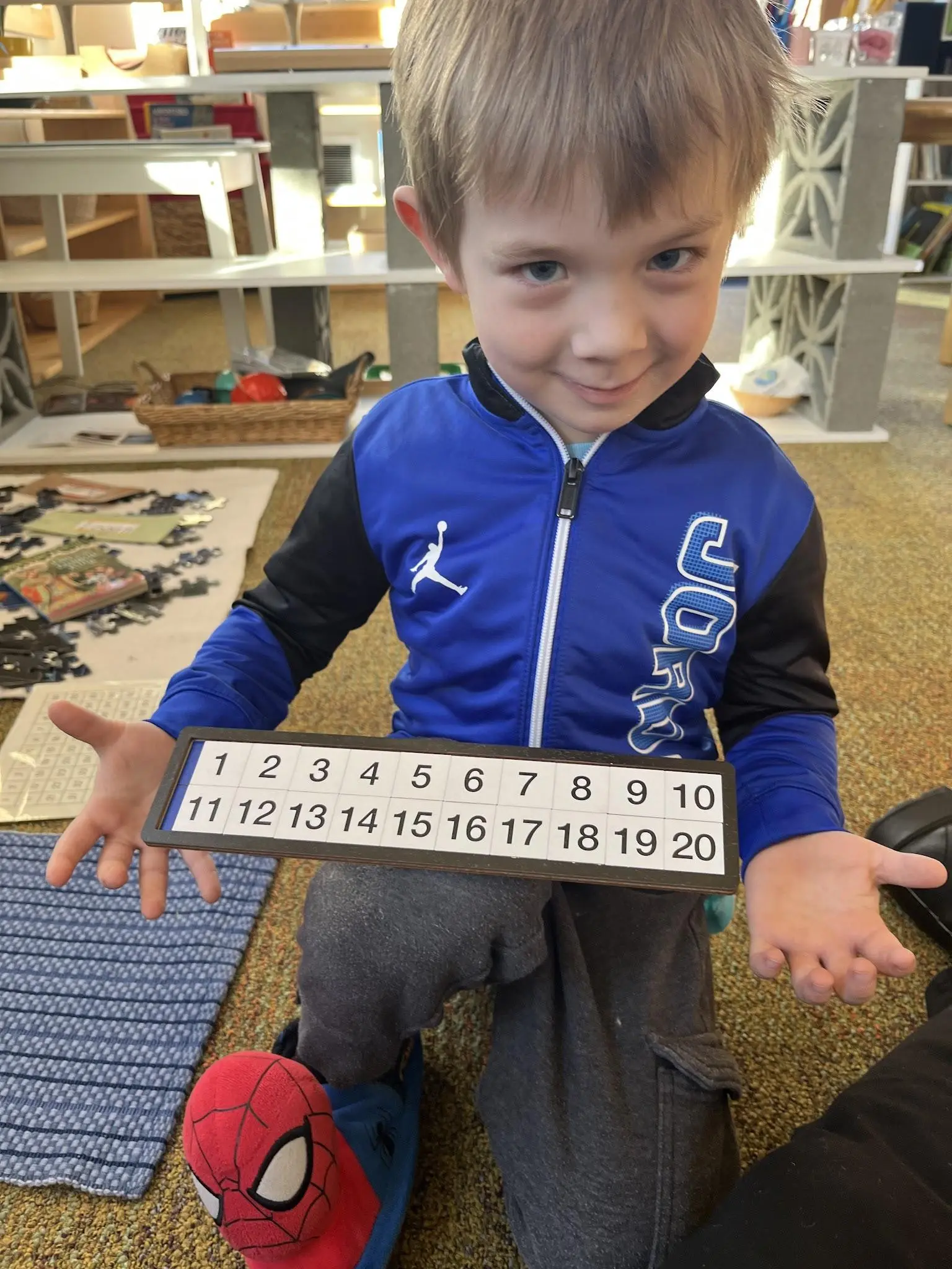 A young boy smiles while holding up a number board in a classroom setting.