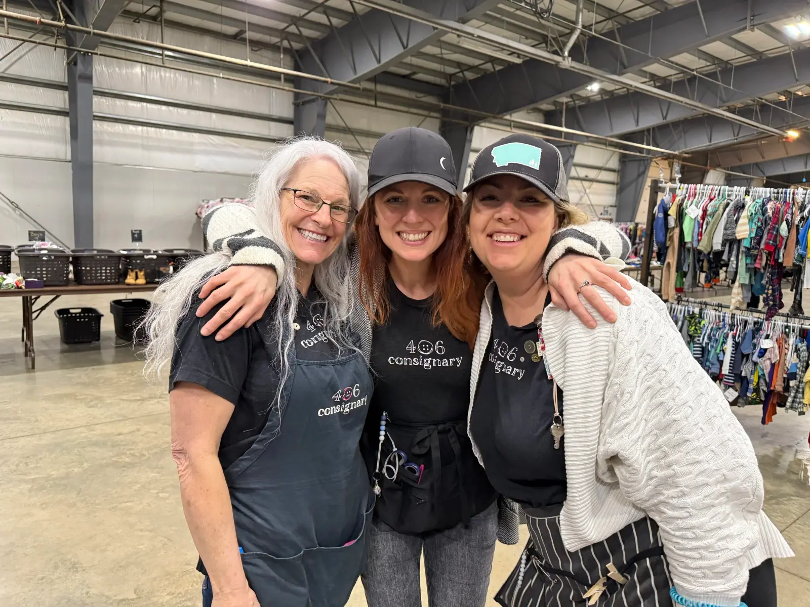 Three smiling women stand with their arms around each other in a large room filled with clothing racks.