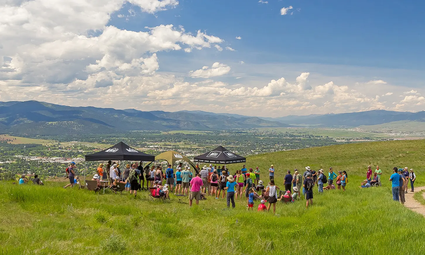 A group of people gathers under tents on a grassy hillside overlooking a valley and mountains.