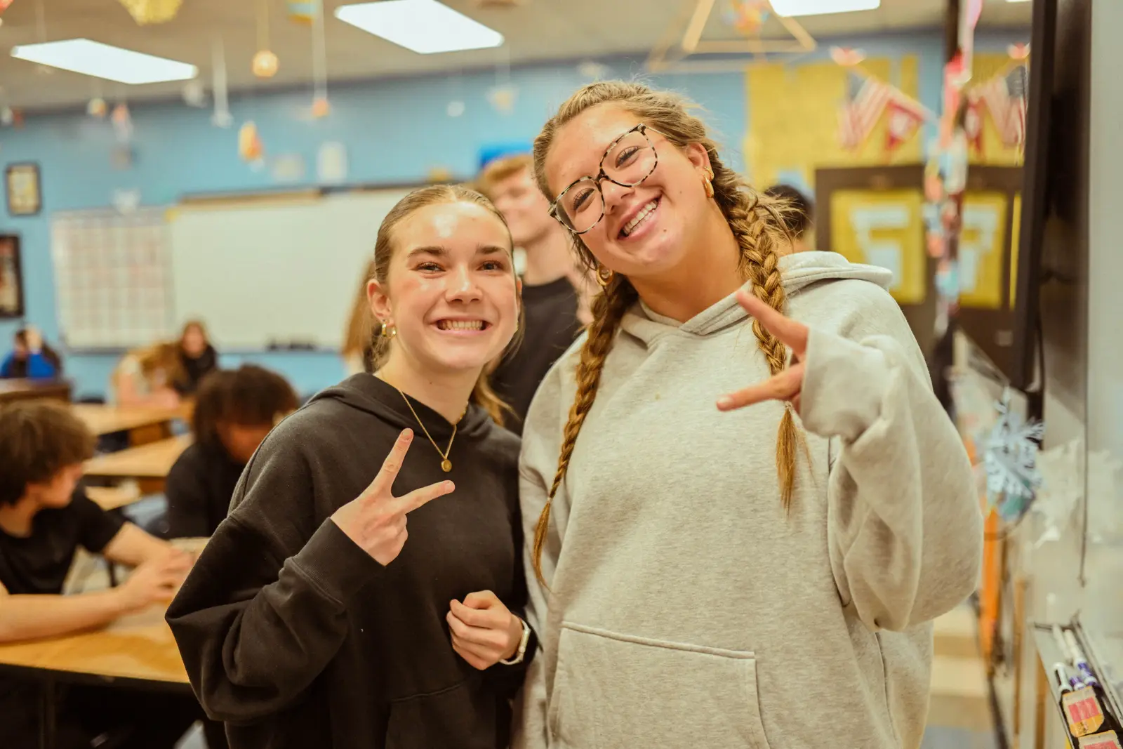 Two young women smile and make peace signs in a classroom.