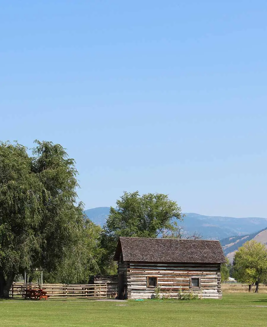 A rustic log cabin sits in a grassy field beneath a clear blue sky with mountains in the background.