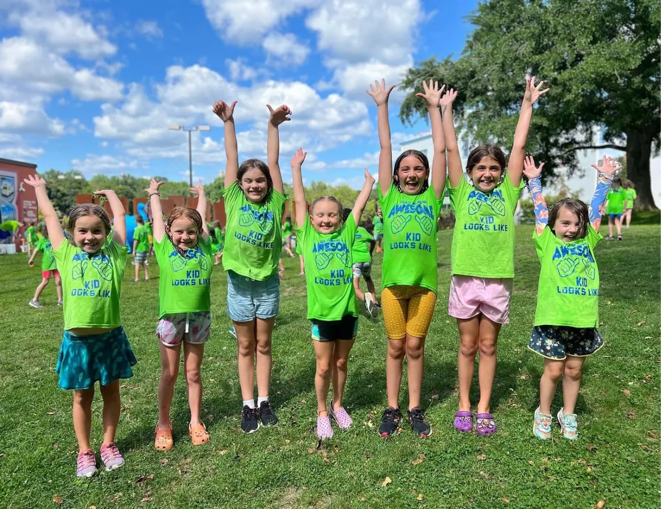 A group of children in matching green shirts are standing in a field with their arms raised.