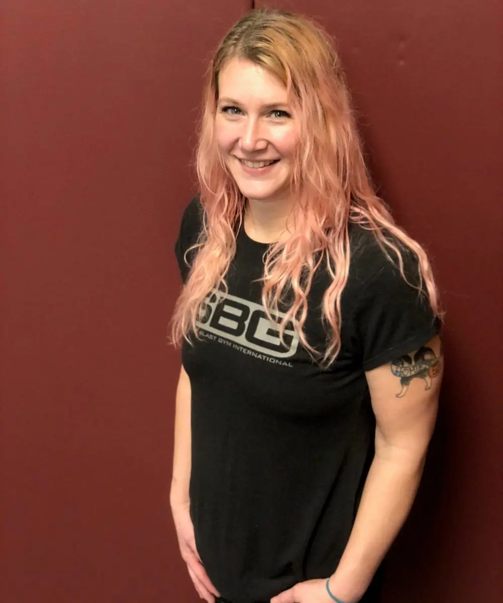 A smiling woman with pink hair wearing a gym t-shirt stands against a maroon wall.