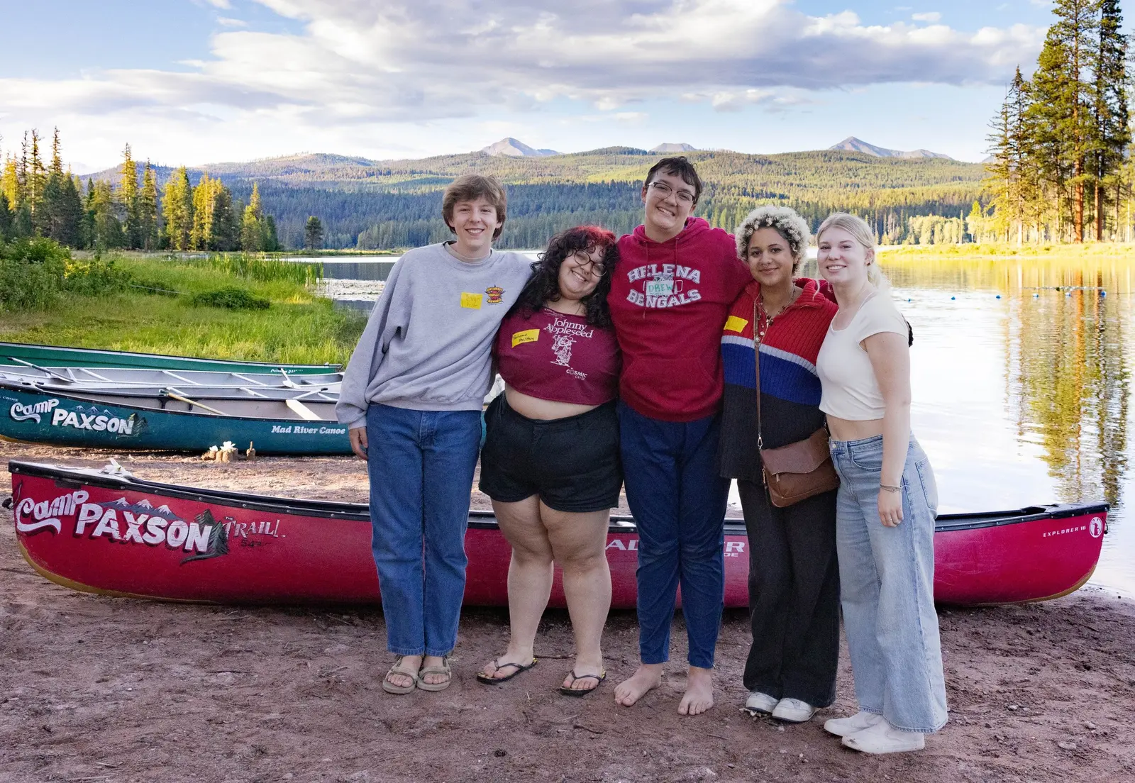 A group of five young people stand together on a lakeshore in front of several canoes.