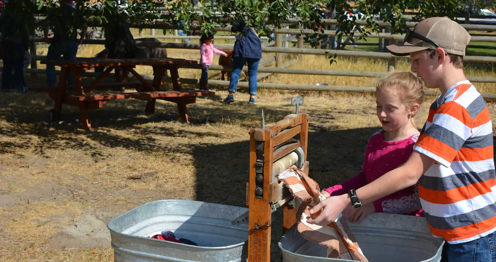 Two children operate a vintage wooden wringer outside in a sunny, rural-style setting.
