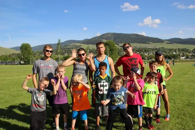 A group of children and young adults pose playfully on a grassy field in front of a mountain landscape.
