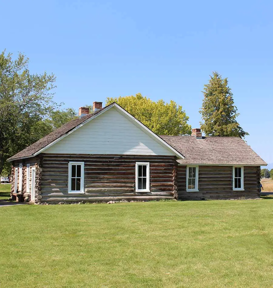 A historic log building sits on a green lawn against a clear blue sky.