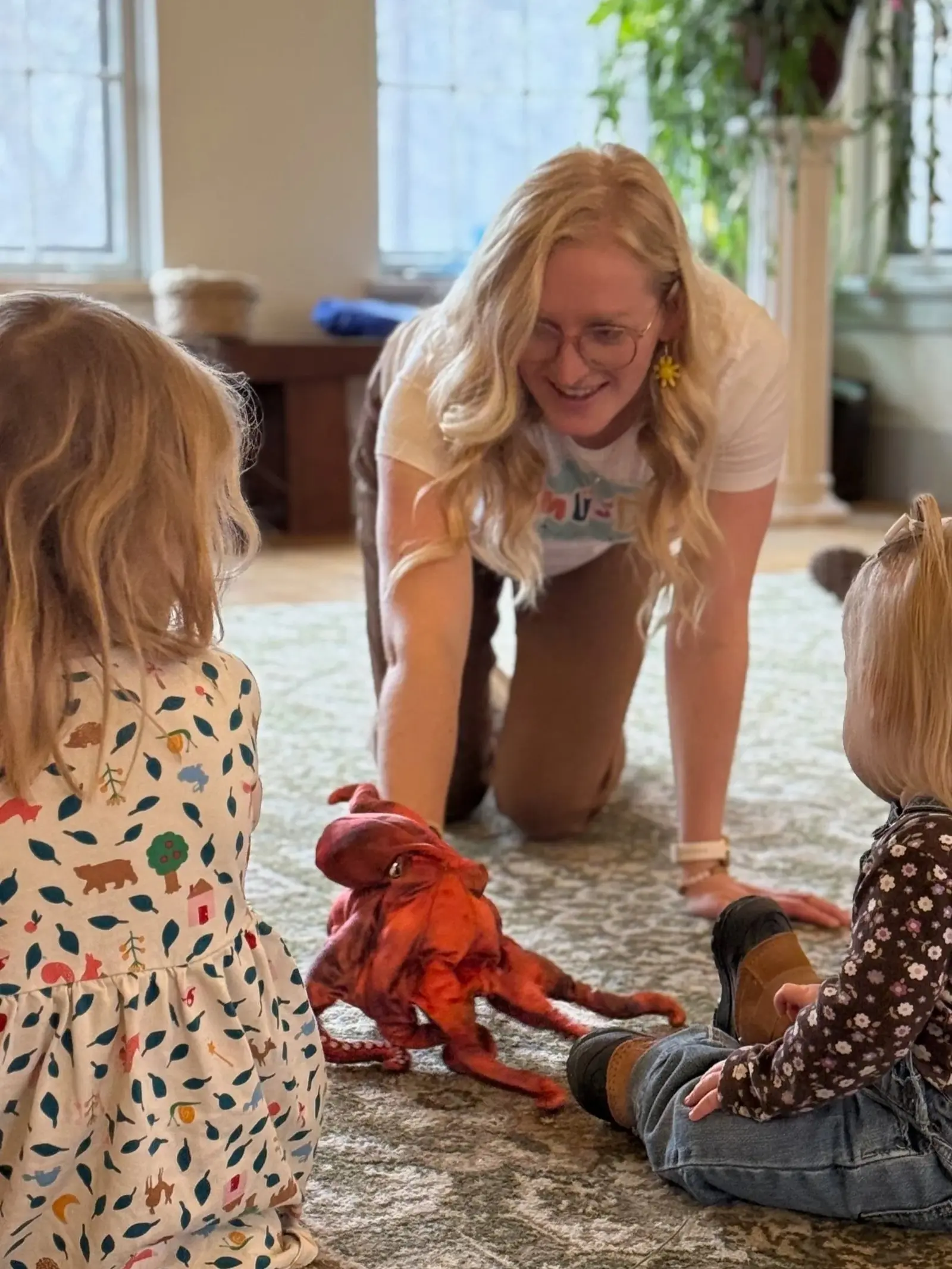 A woman with long blonde hair and glasses engaging with two children sitting on the carpeted floor. One child has her back turned, wearing a dress with colorful animal prints; the other is sitting with her side visible, wearing a floral shirt and jeans. A small red dog is in the center, and they are inside a well-lit room with large windows and green plants in the background.