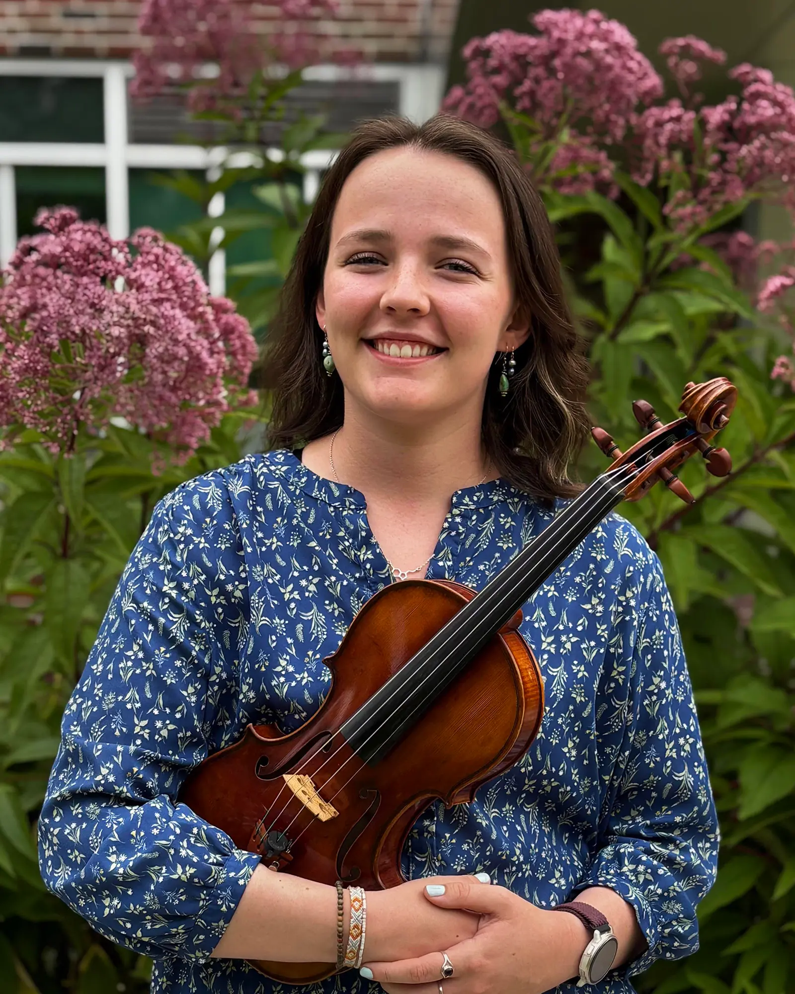 A smiling woman holds a violin while standing in front of flowering bushes.