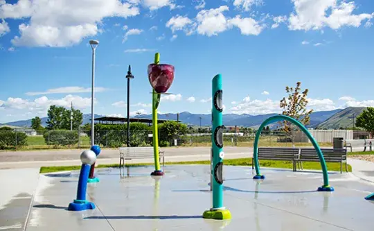 A colorful outdoor splash pad area sits on a concrete surface under a sunny sky.