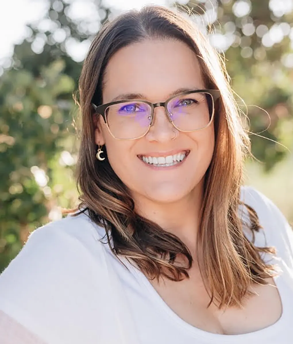 A smiling woman wearing glasses stands outside in natural lighting.
