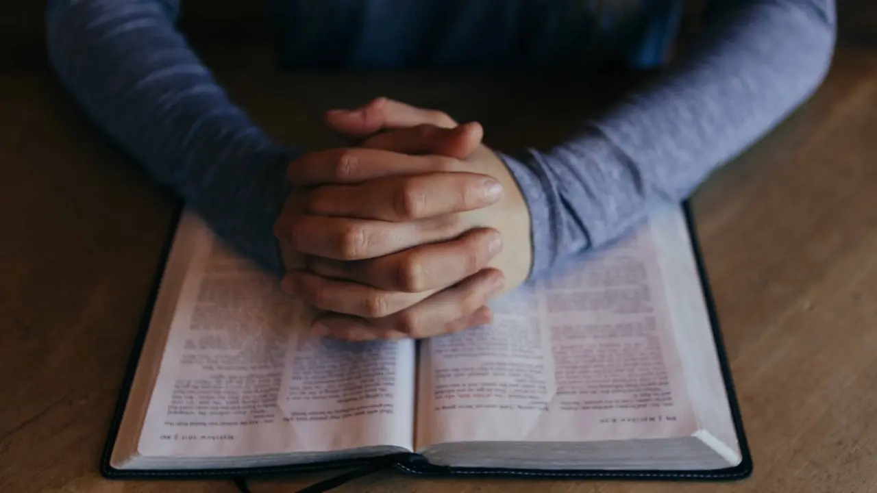 A person has their hands folded in prayer over an open book.