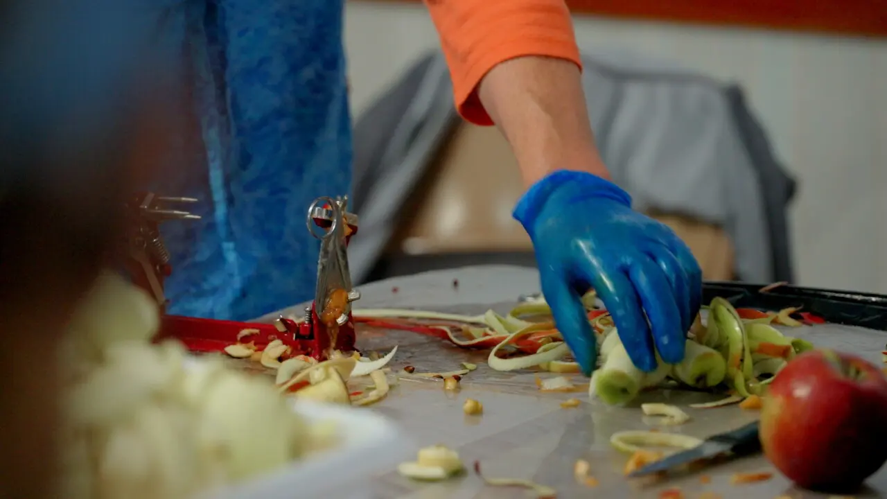 A person wearing a blue glove uses an apple-peeling tool on a table covered with apple pieces and peels.