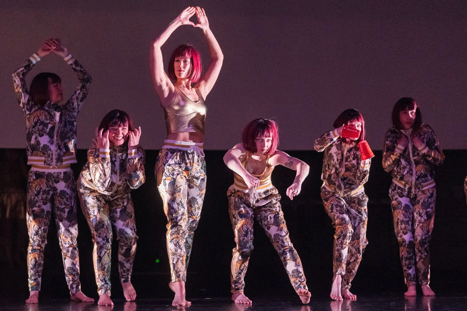 A group of dancers wearing red wigs and matching outfits perform on a stage.