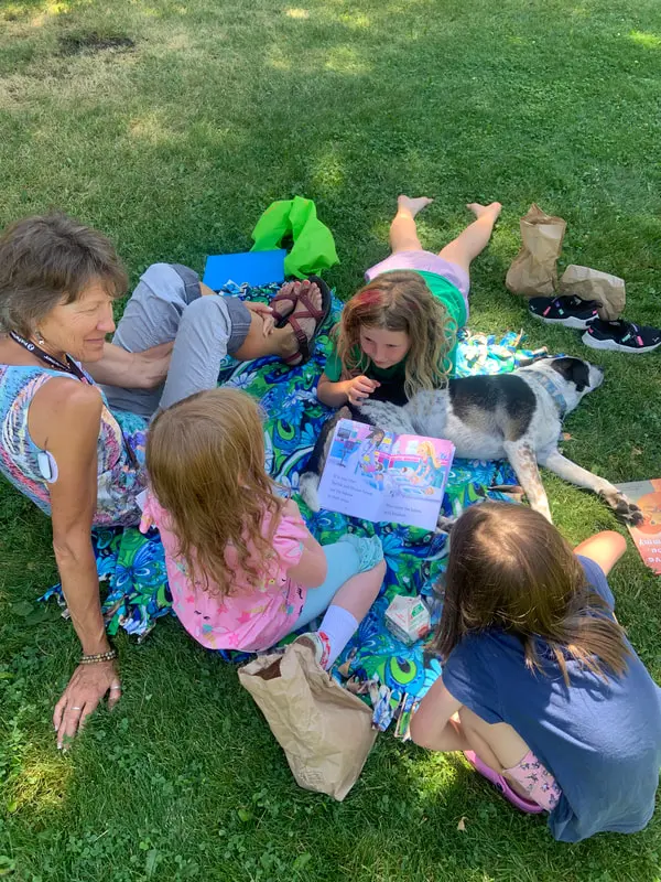 An adult woman and three children read a book together while sitting on a picnic blanket with a dog.