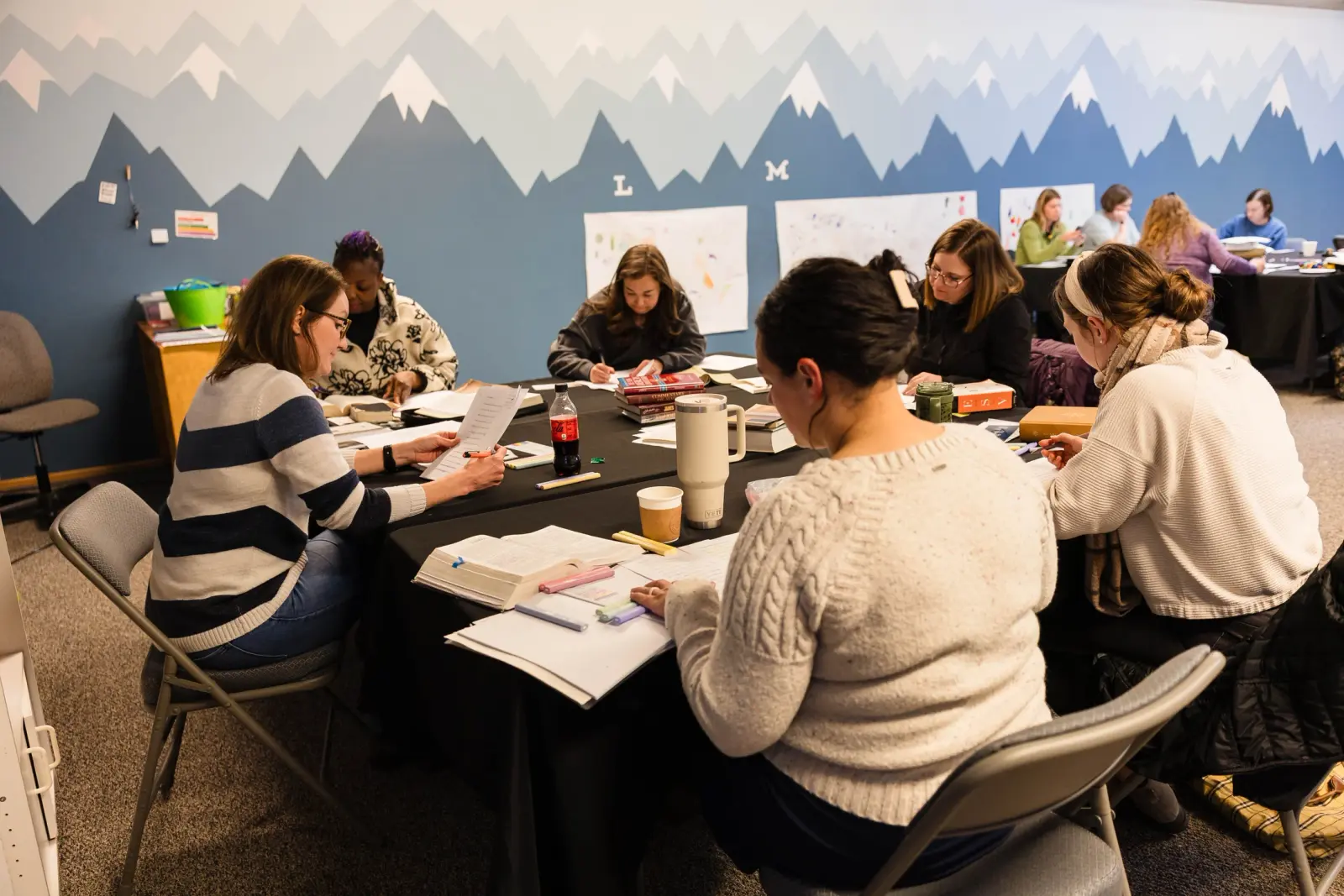A group of women sit around tables studying books and papers in a room with a mountain mural on the wall.