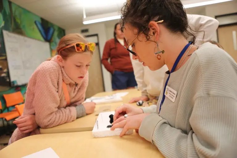 An instructor works with a student on a craft project at a table.