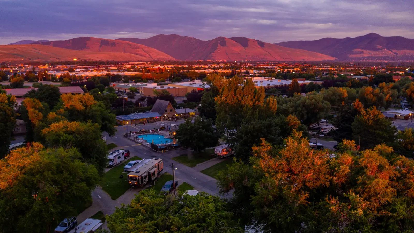 An elevated view of a recreational vehicle park with a swimming pool at sunset, set against a backdrop of distant mountains.