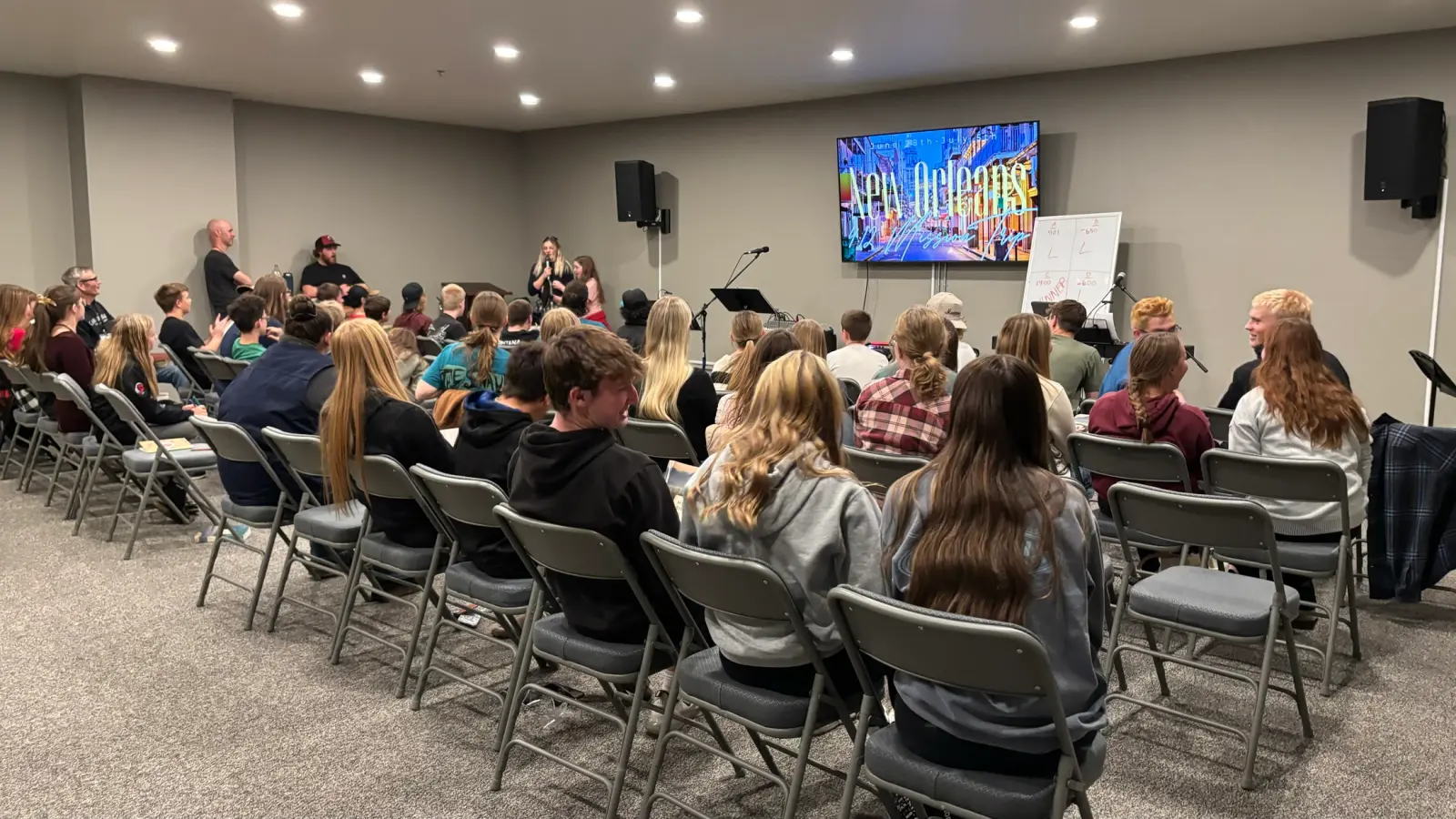 A group of young people sit in rows of chairs listening to a presentation in a meeting room.