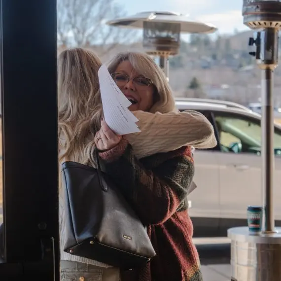 A woman with glasses happily embraces another person while holding papers.