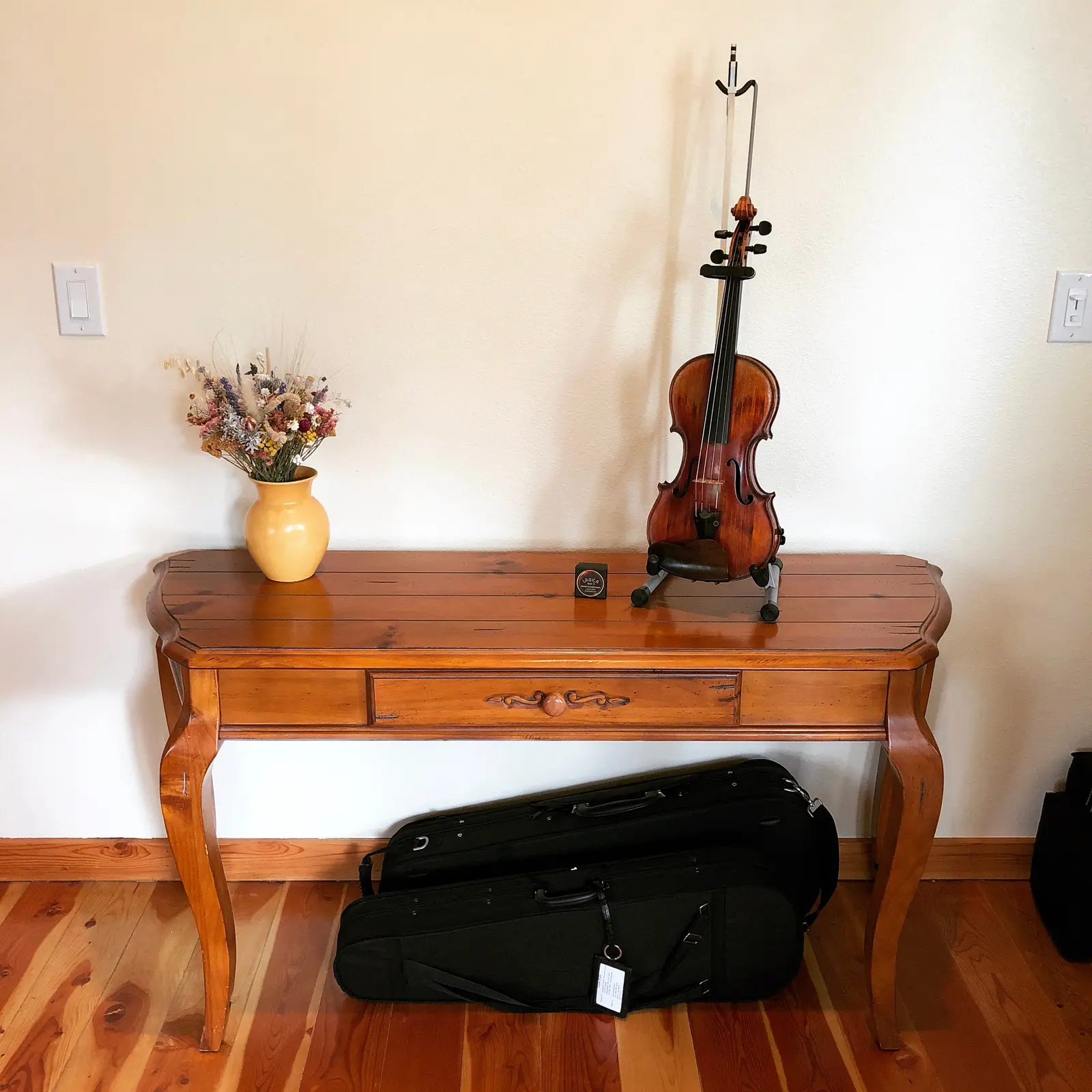 A violin on a stand sits on a wooden table next to a vase of dried flowers.
