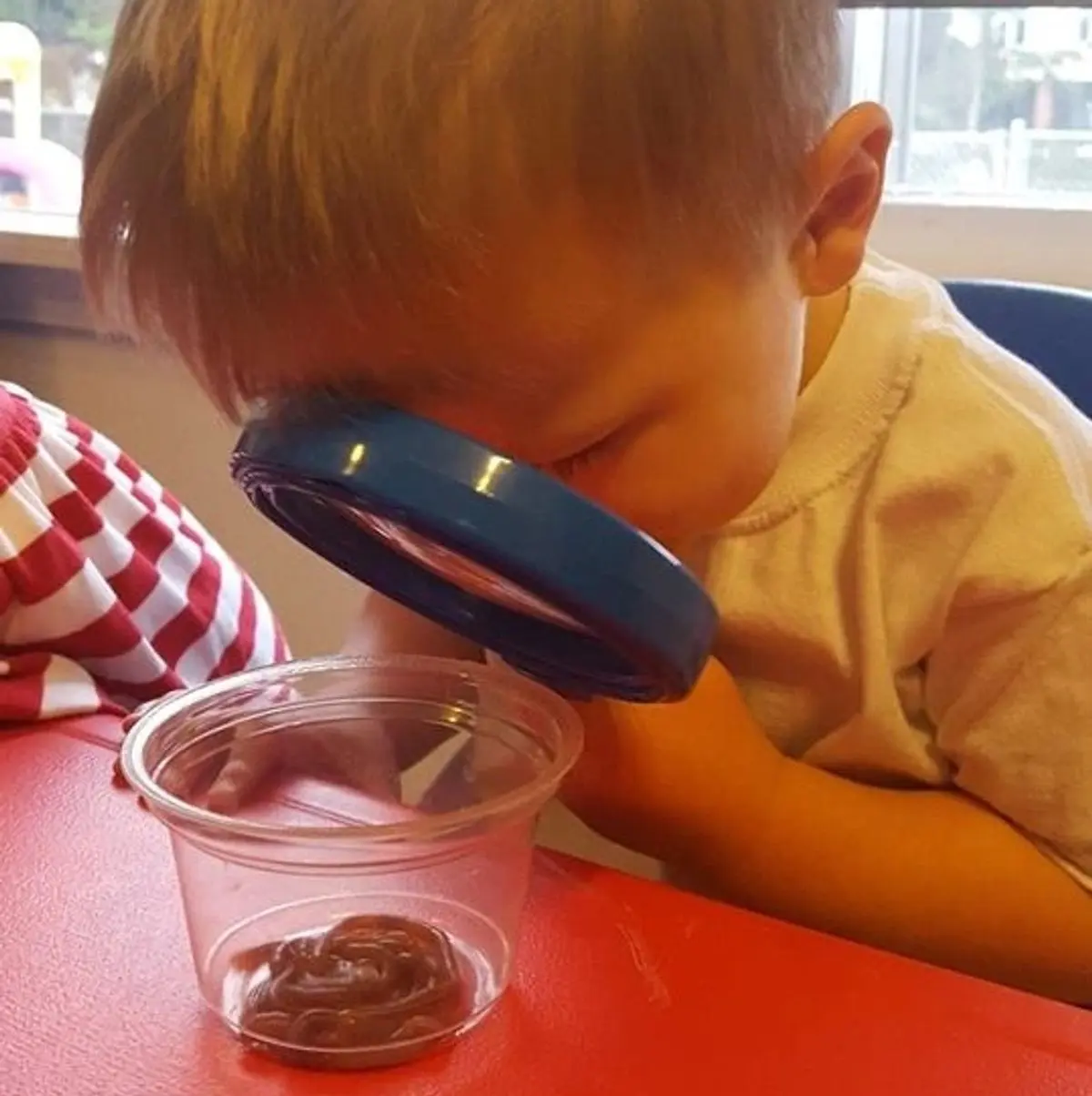 A child uses a magnifying glass to examine a small cup of substance on a red table.