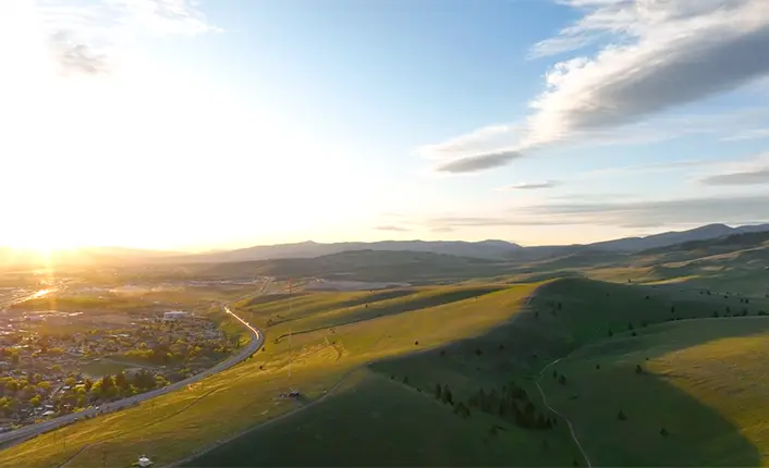 An aerial view shows rolling green hills at sunset near a suburban area.