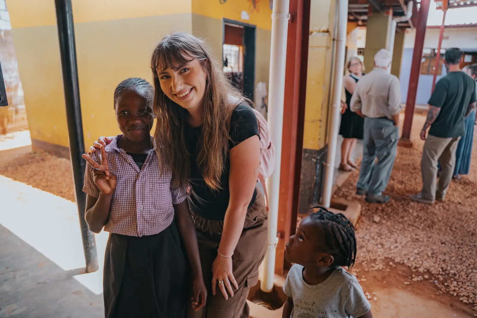 A woman smiles while posing with a young student who makes a peace sign, with another young child looking up at them nearby.