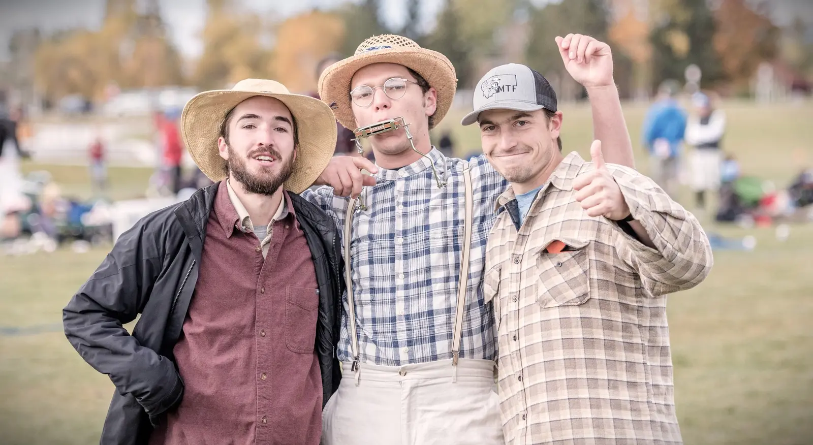 Three men pose together outdoors, with one wearing a hat and playing a harmonica.