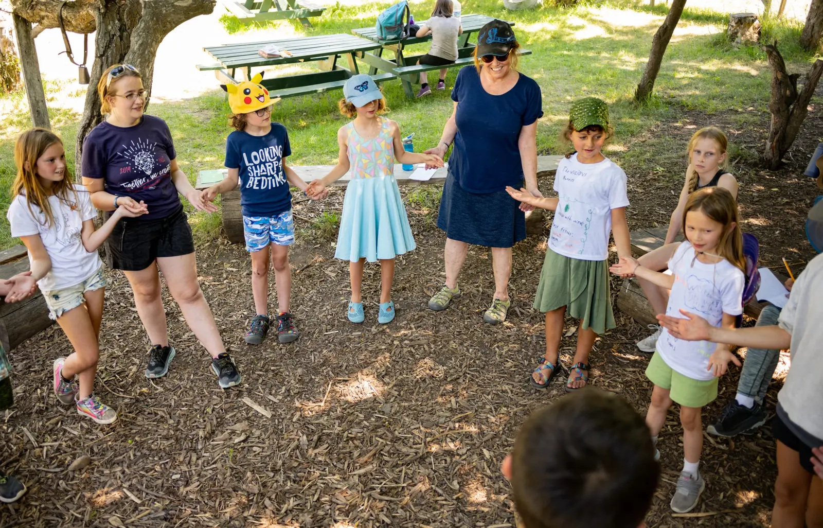 A group of children and adults stand in a circle outdoors holding hands.