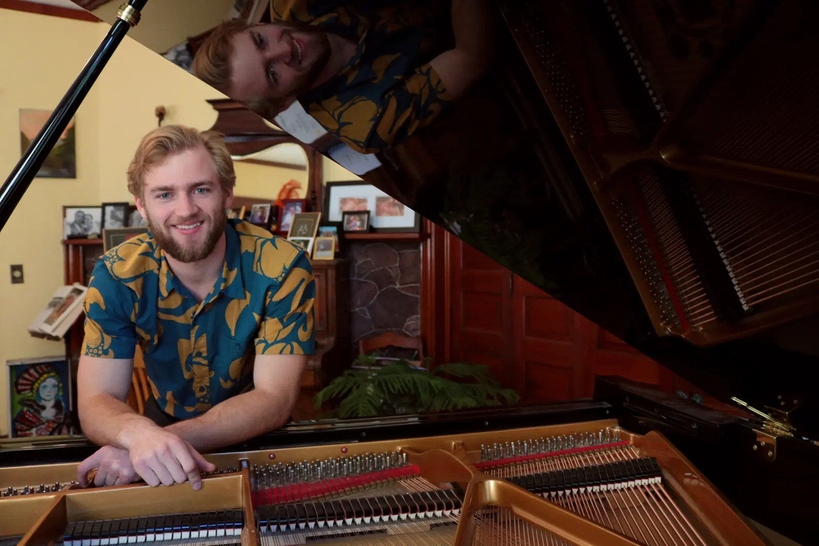 A man in a patterned shirt smiles while leaning on the open lid of a grand piano.