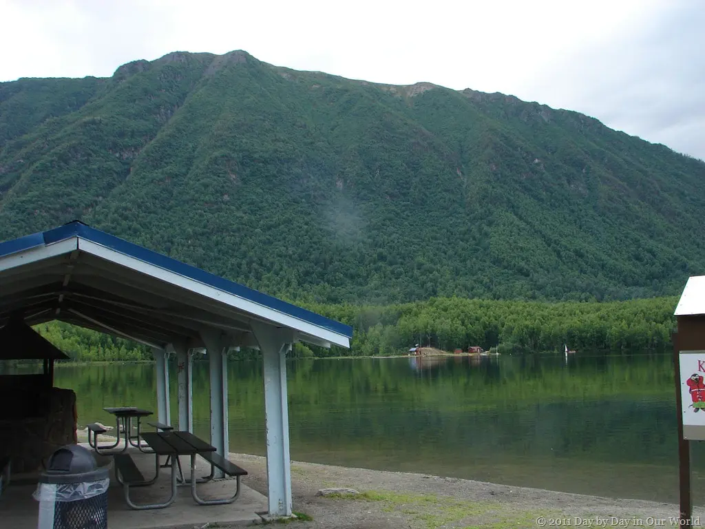 A picnic shelter sits on the shore of a lake with a large mountain in the background.