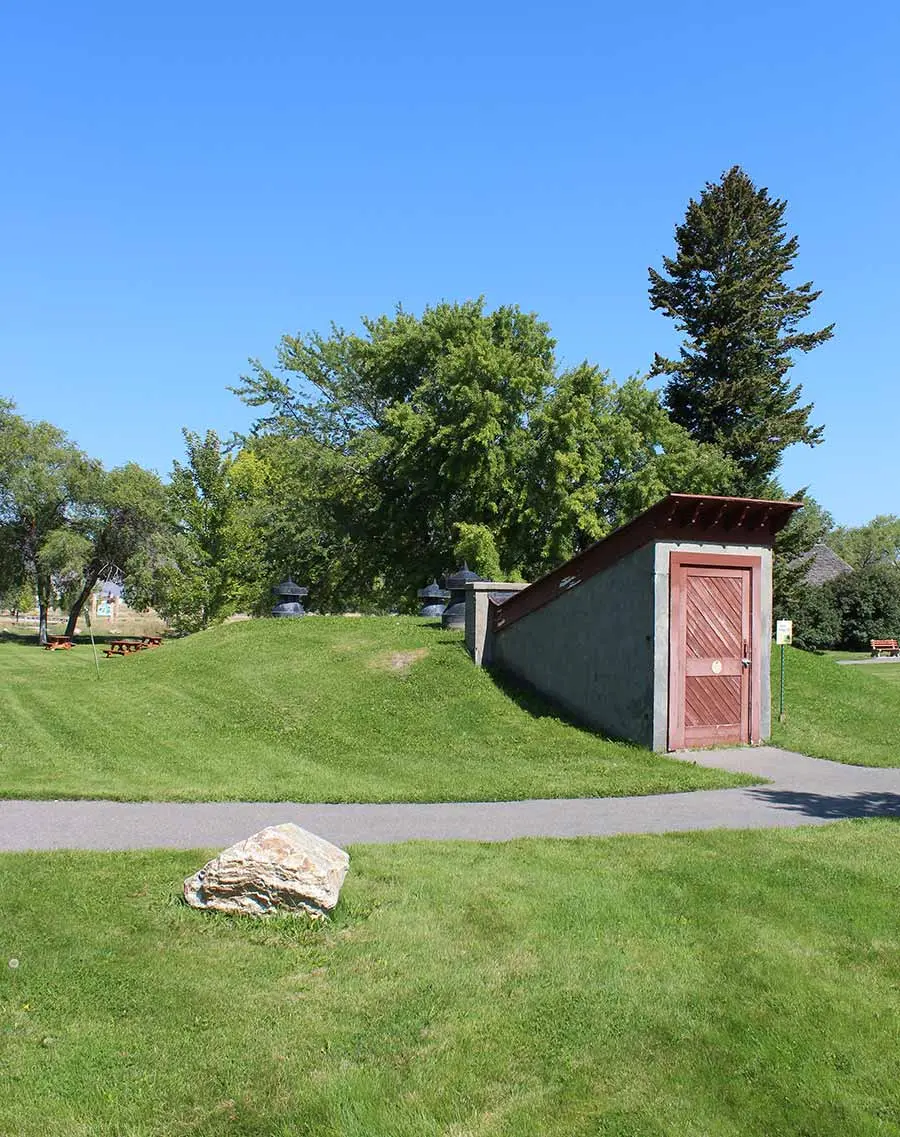 An earthen cellar entrance sits in a grassy park setting under a clear blue sky.