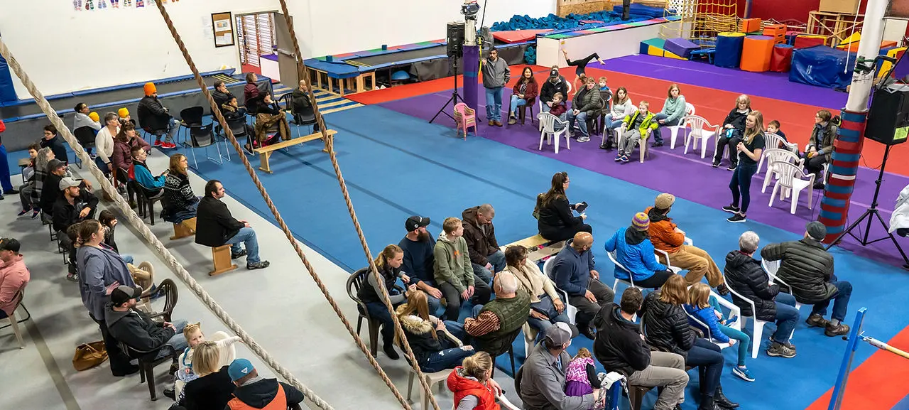 A large group of people are gathered on mats in a gym, watching an activity.