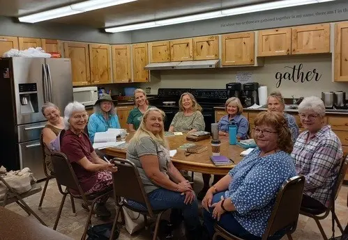 A group of women sit around a wooden table in a kitchen area.