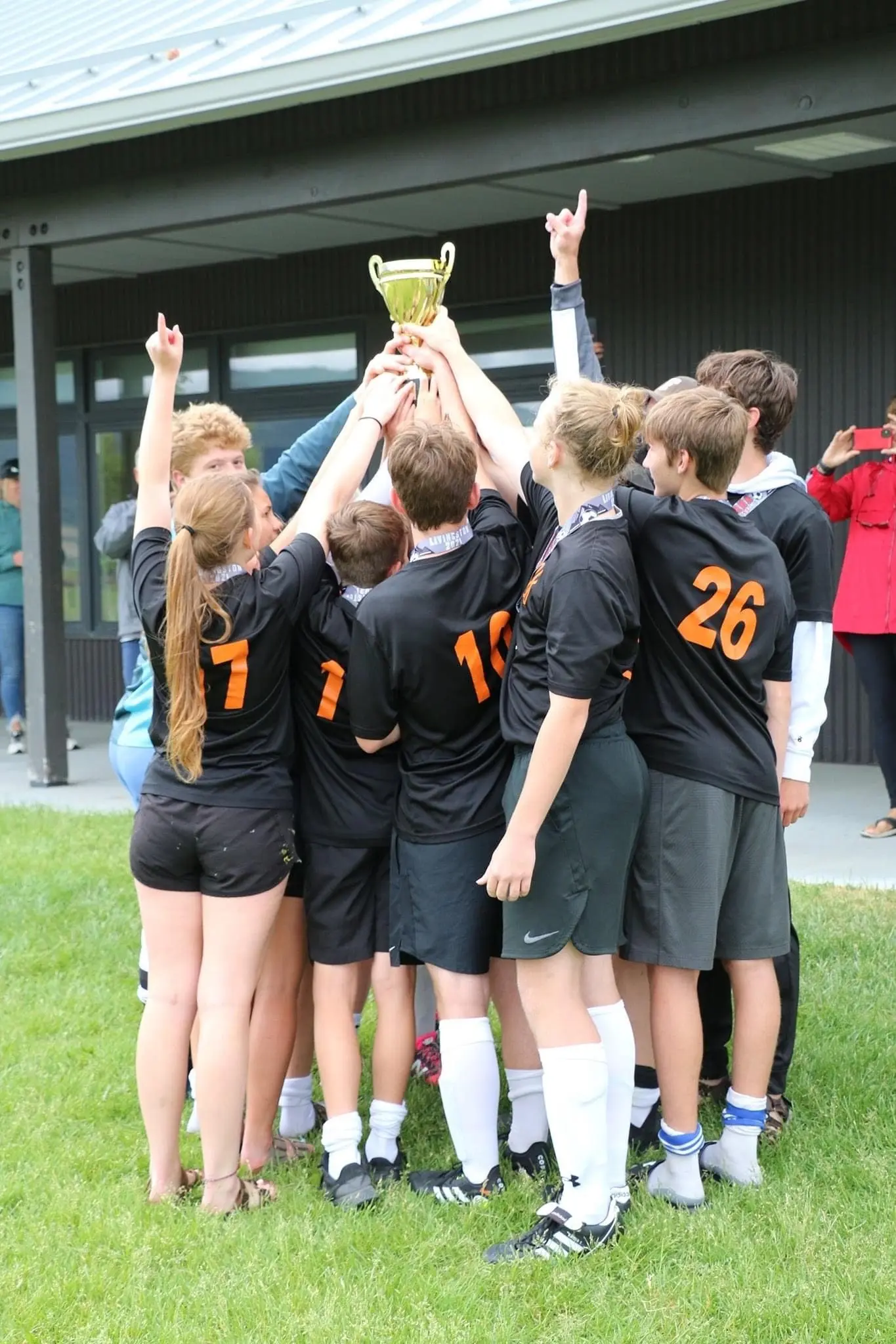 A team of young athletes in black and orange jerseys lifts a trophy in celebration.
