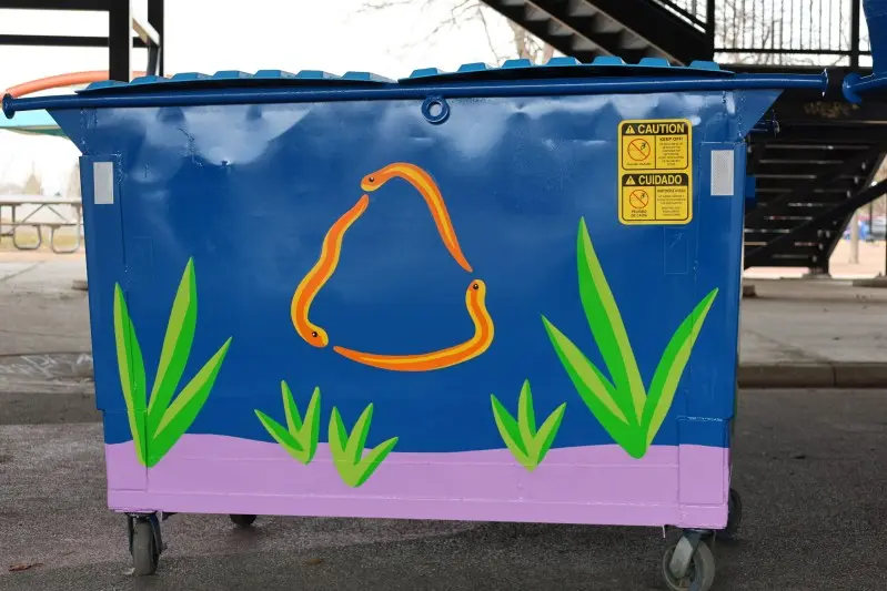 A blue dumpster is painted with images of snakes forming a triangle and green plants.