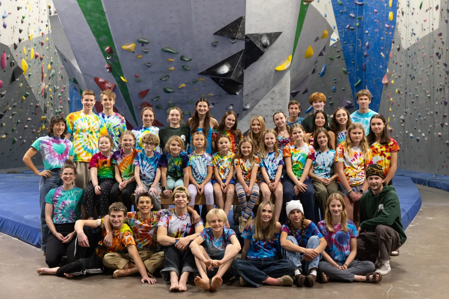 A large group of young people wearing tie-dye shirts poses together in front of a climbing wall.