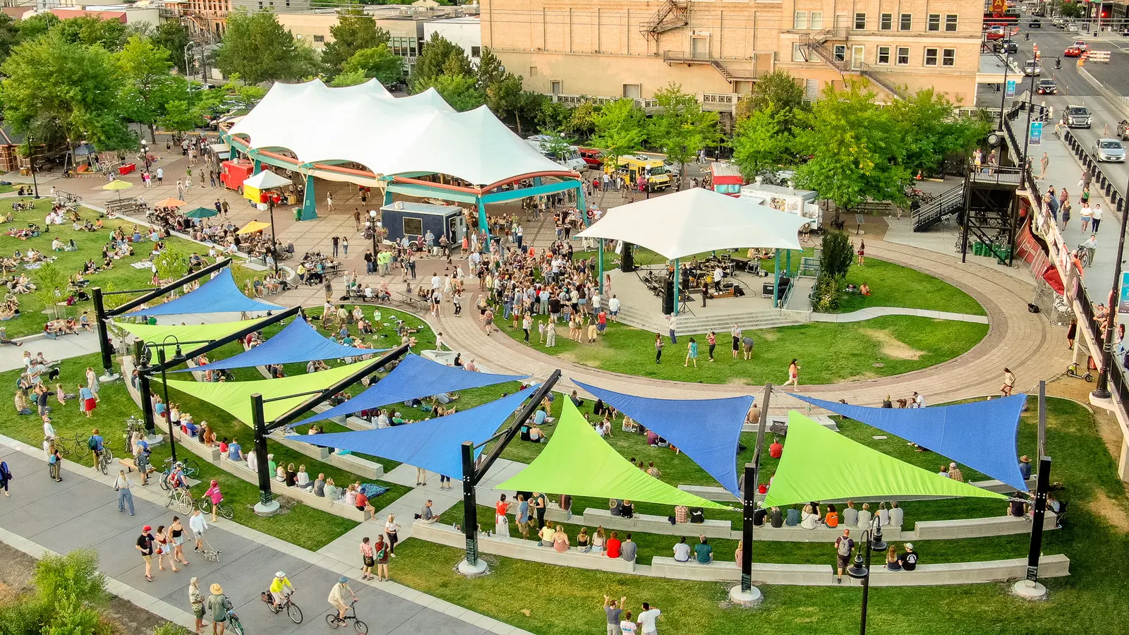 A high-angle view shows people gathering in a park area with a stage and shade sails.