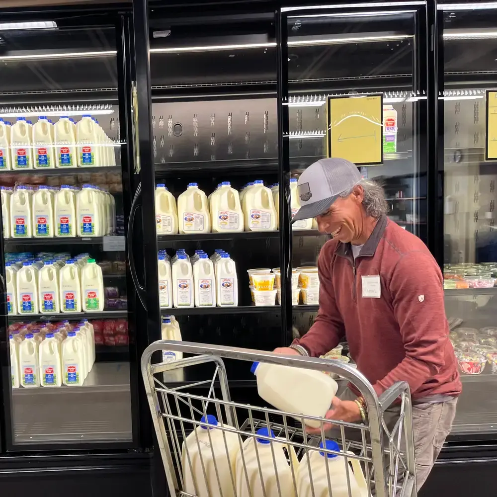 A man wearing a cap and long-sleeved shirt laughs while placing a gallon of milk into a shopping cart in front of refrigerated dairy cases.