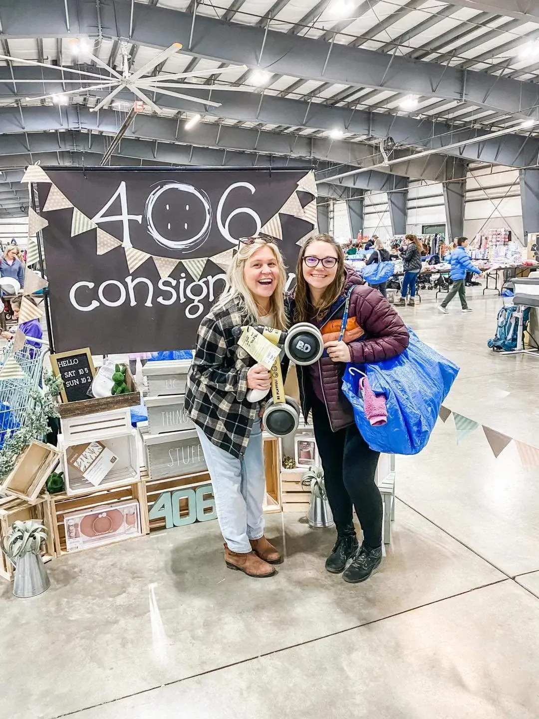 Two women pose for a photo in front of a consignment sale booth at an event.