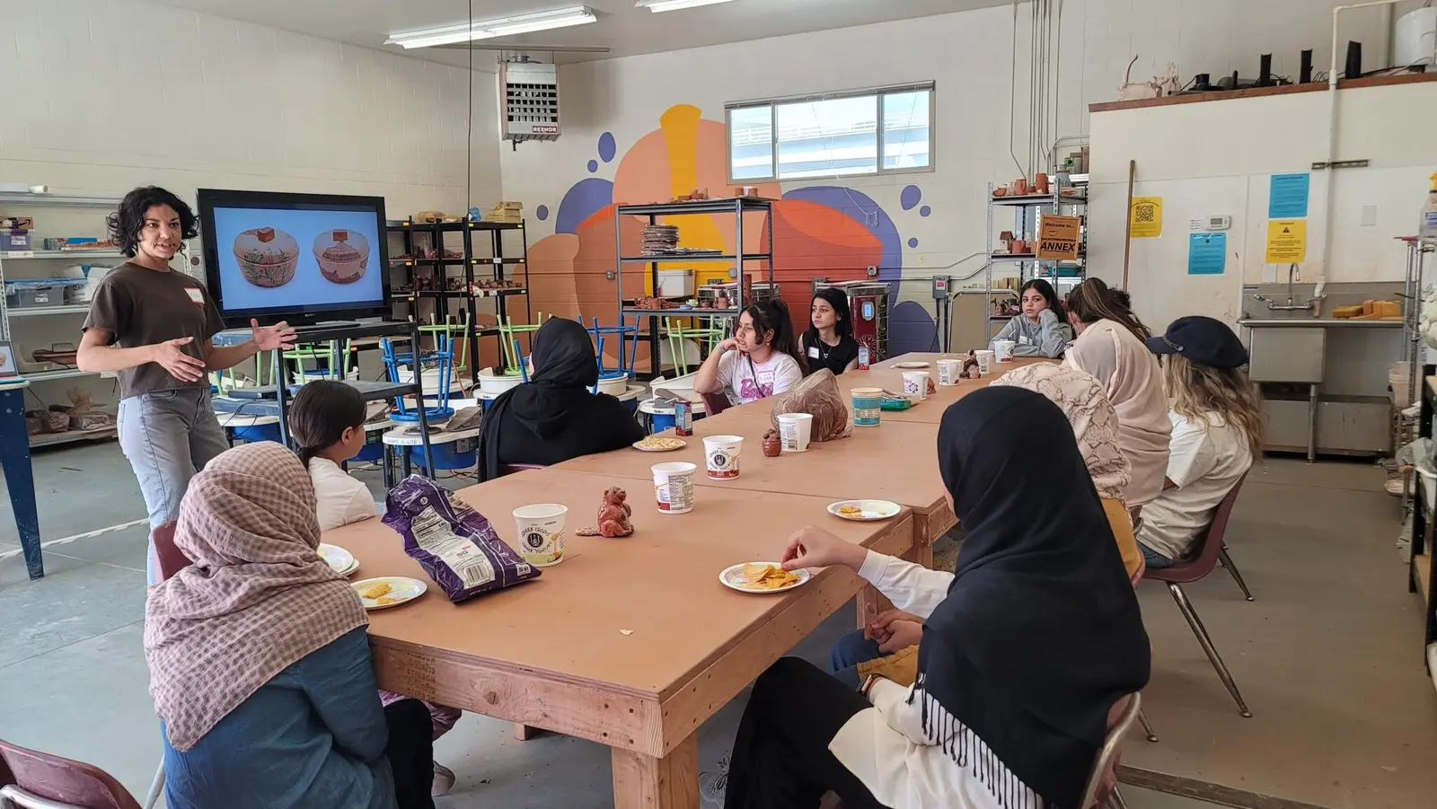 An instructor gives a presentation to a group of young women gathered around a large table in a pottery studio.