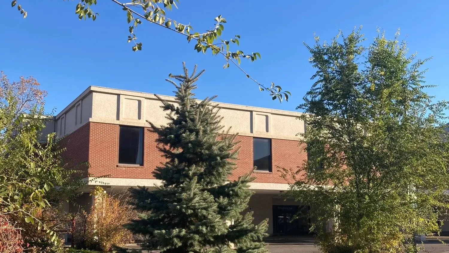 A two-story building made of brick and tan stucco is framed by trees against a clear blue sky.