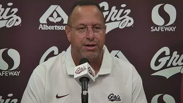 A man with glasses speaks into a microphone in front of a branded backdrop.