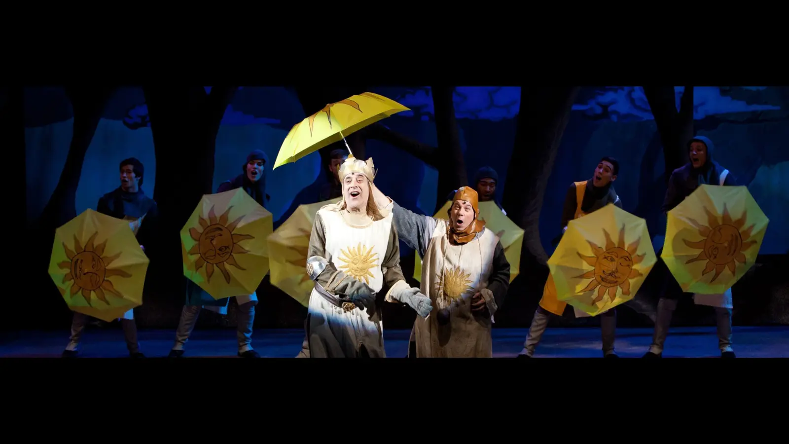 A cast of actors in costume performs on stage holding yellow sun-themed umbrellas.